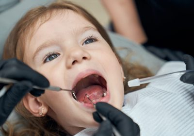 Portrait of pretty bold female child sitting in dental chair with open mouth while professional doctor in rubber gloves carefully examining condition of teeth. Concept of dentistry